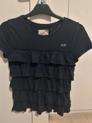 Mörkblå volangtopp från Hollister - Supersöt mörkblå topp från Hollister med flera lager volanger framtill och liten broderad logga på bröstet. Kortärmad modell i bomull