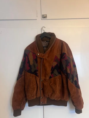 Brun vintage skinnjacka med mönster - Unik brun skinnjacka i vintage-stil med färgglada mönstrade partier i vinrött, mörkblått och olivgrönt på ärmar och axlar. Jackan har breda muddar, tryckknappar och en cool krage med spänne. Perfekt för dig som vill sticka ut med retrovibbar. Finns ett litet hål på ryggen, men det märks inte av. Märket är Vera Pelle 