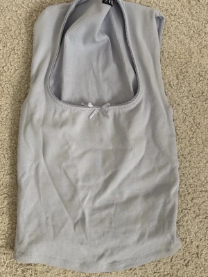 Ljusblå ribbad linnetopp från Zara - Snygg ljusgrå ribbad linnetopp från Zara med djup rundad urringning och en liten rosett framtill. Toppen är ärmlös och har en mjuk, stretchig känsla som passar perfekt till jeans eller kjol.