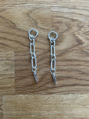 Silvriga kedjeörhängen med spetsdetalj - Säljer ett par coola örhängen i silverfärgad metall med kedjor och spetsiga hängen längst ner. Örhängena har en modern och edgy vibe med sina avlånga länkar och minimalistiska design. Perfekta för dig som gillar statement-accessoarer.