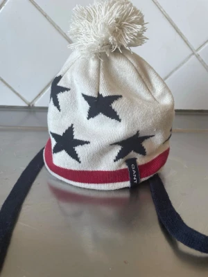 Vit mössa med stjärnor från Gant - Säljer en vit mössa från Gant med svarta stjärnor, röd rand och mörkblå knytband. Mössan har en stor tofs på toppen och är tillverkad i mjukt stickat material. Perfekt för dig som vill ha en snygg och bekväm accessoar till vintern. Barnmössa