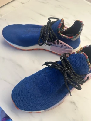 Adidas Pharrell Williams blå sneakers - Säljer ett par Adidas Pharrell Williams sneakers med blå ovandel i mesh, rosa detaljer på sidorna och färgglad kant runt öppningen. Skorna har svartvita snören och vit sula med orange yttersula. Perfekta för dig som gillar unika och färgstarka sneakers.