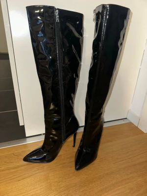 Svarta lackade knähöga stövlar - Säljer ett par svarta, glansiga knähöga stövlar med spetsig tå och hög smal klack. Stövlarna har dragkedja på insidan. UK size 5 (37-38)