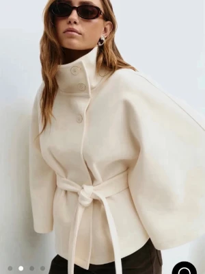 Krämvit kappa med bälte och hög krage - Helt ny. Stilren krämvit kappa med breda ärmar, hög krage med knappar och knytskärp i midjan. Kappan har en modern, boxig siluett och är tillverkad i ett tjockare tyg som ger struktur. Perfekt för dig som gillar minimalistisk och trendig design.