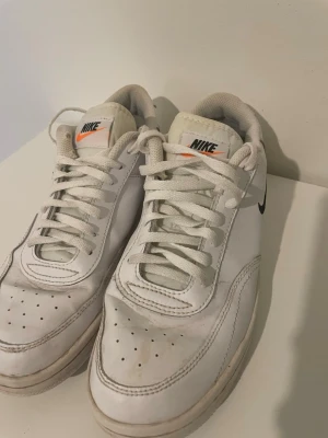 Vita Nike Air sneakers med svart swoosh - Fräscha vita Nike Air sneakers med klassisk svart swoosh på sidan och orange detalj på tungan. Skorna har lågt skaft, perforerad tå och är tillverkade i läder med textilfoder. Perfekta för en clean och sportig look, men har tappat bort en av sulorna.