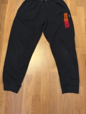 Svarta mjukisbyxor från Hoodrich - Säljer ett par svarta mjukisbyxor från Hoodrich med broderad logga i orange och rött på ena benet. Byxorna har resår i midjan och vid bensluten för en skön och avslappnad passform. Perfekta för chill eller streetwear-stil.