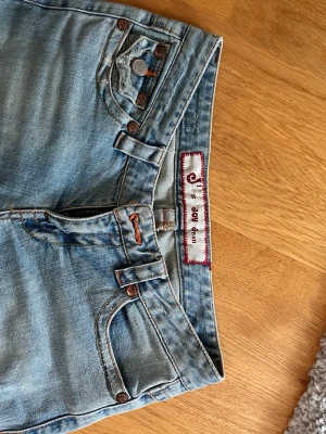 Ljusblå slitna skinny jeans J-LO - Säljer ett par ljusblå bootcut jeans från J-LO by Jennifer Lopez med slitningar på låren och klassisk femficksdesign. Jeansen har dragkedjegylf, bälteshällor och är tillverkade i denim med stretch för en bekväm passform. Sitter perfekt på mig som vanligtvis har strl 36