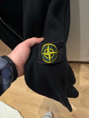 Svart ziptröja från Stone Island - Svart ribbstickad ziptröja från Stone Island med klassisk logotyp-patch på ärmen. Tröjan har hög krage och dragkedja framtill, perfekt för lager-på-lager. Materialet är grovstickat och ger en stilren och sportig look.