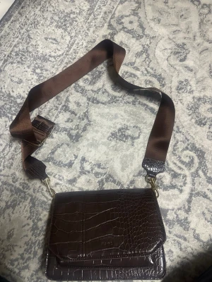 Brun axelväska i krokopräglat läder - Snygg brun axelväska med krokodilpräglat mönster och bred justerbar axelrem i textil. Väskan har guldfärgade detaljer och praktiskt magnetlås. Pris kan diskuteras!