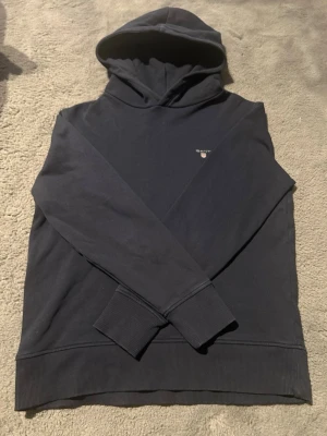 Mörkblå hoodie från GANT - Snygg mörkblå hoodie från GANT med liten broderad logga på bröstet. Klassisk modell med huva och ribbade muddar vid ärmslut och nederkant. Tillverkad i mjukt bomullsmaterial som känns skönt mot huden. Perfekt för en avslappnad och stilren look.