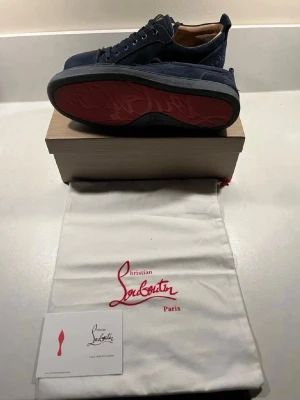 Christian Louboutin - Snygga mörkblå sneakers från Christian Louboutin med ikonisk röd sula och lågt skaft. Ovandelen är i mocka och insidan har en beige ton. Skorna har klassisk snöring och skorna kommer med allt og 