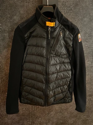 Svart vindjacka från Parajumpers youth stlk M - Svart vindjacka från Parajumpers med quiltad front och mesh-liknande ärmar. Jackan har dragkedja framtill, bröstficka med dragkedja och en gul detalj vid kragen. Klassisk PJS-patch på ärmen ger en sportig vibe. Perfekt för dig som gillar stilren och funktionell design.