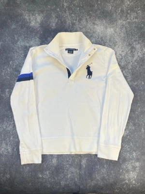 Vit långärmad tröja från Ralph Lauren - Vit långärmad polotopp från Ralph Lauren med blått broderat logotyptryck på bröstet och en blå rand på ena ärmen. Toppen har hög krage med knappar och är gjord i mjuk bomull. Perfekt för en sportig och stilren look.