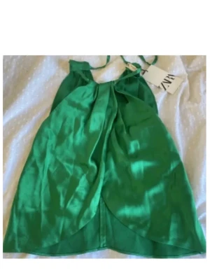 Grön satinlinnetopp från Zara - Snygg grön linnetopp från Zara i glansigt satinmaterial. Toppen har tunna axelband och en lös, flowy passform med omlottdetalj framtill. Perfekt för dig som vill sticka ut med en färgstark och trendig topp.