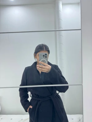 Svart kappa med knytskärp - Stilren svart kappa med knytskärp i midjan och bred krage. Medel lång modell med fickor på sidorna, perfekt för kyligare dagar. Klassisk design som passar till många olika outfits.