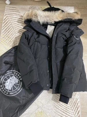 Svart parkas från Canada Goose - Snygg svart parkas från Canada Goose med avtagbar huva och äkta pälsdetalj. Jackan har klassisk logotyp på ärmen, dragkedja och knappar framtill samt ribbade muddar. Perfekt för kalla vinterdagar och riktigt varm.
