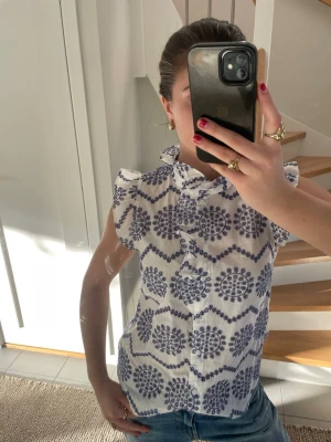 Vit/blå blus med volanger - Snygg vit blus från Holly & Whyte med blått mönster. Blusen är ärmlös med volangdetaljer vid axlarna. Från Holly & Whyte. 