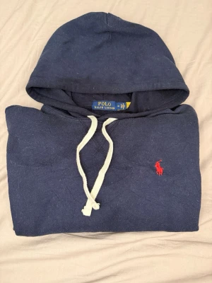 Ralph lauren hoodie mörkblå - En riktig klassiker.  Mörkblå hoodie från Polo Ralph Lauren med broderad röd logga på bröstet. Clean, tidlös och lätt att styla – funkar lika bra till vardags som till en mer uppklädd look.  • 100% original Polo Ralph Lauren   • Storlek: M • Färg: Navy   • Skick: Bra – normalt bruksspår   • Mjuk och bekväm kvalitet   