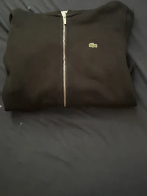 Svart zip-hoodie från Lacoste - Svart hoodie med dragkedja från Lacoste, klassisk krokodil-logga på bröstet. Mjuk bomullskänsla, ribbade muddar och två fickor framtill. Perfekt för en chill och stilren look.