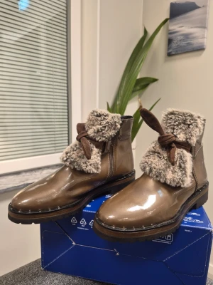 Marc Bruna lackade stövletter med päls 40 - Snygga bruna stövletter med glansig lackyta och mjuk beige fuskpäls runt skaftet. Dekorativa bruna snören framtill och grov sula för extra grepp. Dragkedja på sidan för enkel påtagning. Perfekta för dig som vill ha både stil och värme.