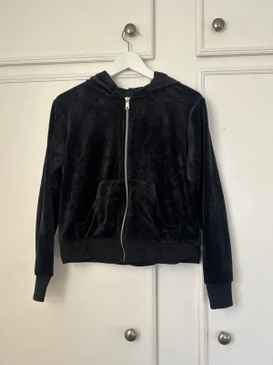 Svart velourhoodie med dragkedja - Mjuk och skön svart hoodie i velour med dragkedja framtill och kängurufickor. Klassisk huva och ribbade muddar vid ärmslut och nederkant. Perfekt för en chill och avslappnad stil.