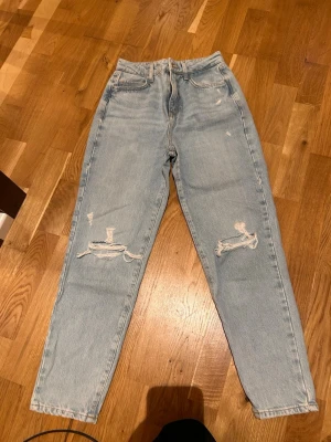 Ljusa mom jeans med slitningar - Säljer ett par ljusblå mom jeans med hög midja och trendiga slitningar på båda knäna. Klassisk femficksmodell i mjukt denimtyg. Perfekta för en avslappnad och cool stil.