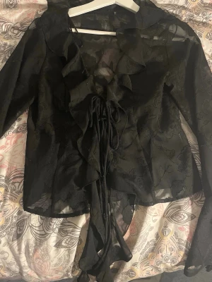 Svart transparent blus med volanger - Snygg svart blus i transparent tyg med subtilt blommönster. Blusen har långa ärmar, volangdetaljer längs framkanten och snörning framtill. Perfekt för dig som gillar en edgy och romantisk stil. Materialet känns lätt och luftig.