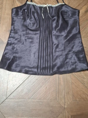 Svart linnetopp med snörning - Snygg svart linnetopp i satin med tunna axelband och dekorativ snörning framtill. Toppen har veckade detaljer längs mitten och en lätt glansig finish som ger en lyxig känsla. Perfekt att styla med jeans eller kjol.