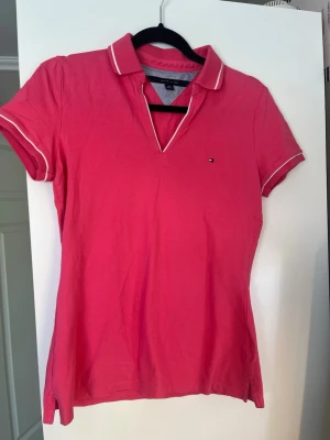 Rosa pikétopp från Tommy Hilfiger - ÄKTA!! Snygg rosa pikétopp från Tommy Hilfiger med vit kant på krage och ärmslut. Klassisk polokrage och liten logga på bröstet. Tillverkad i bomull för en bekväm känsla och perfekt passform. Inga tvättlappar kvar, och tyvärr för liten så har ingen bild på. Passar XS/S