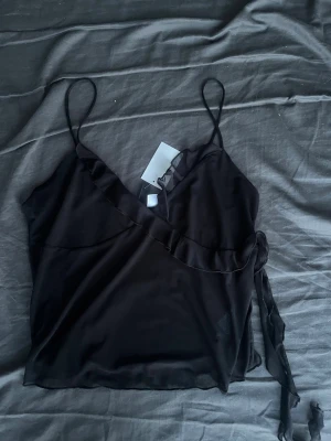 Svart omlott linnetopp med volang - Snygg svart omlott linnetopp med tunna axelband och volangdetalj längs ringningen. Toppen har knyt i sidan och är gjord i ett tunt, lätt material som känns luftigt. Perfekt för dig som gillar en enkel men trendig look.