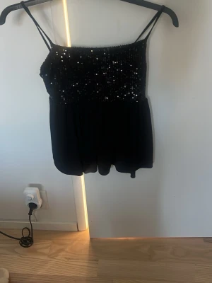 Svart paljettlinnetopp med tunna band - Snygg svart linne-topp med tunna axelband och paljetter upptill som ger ett glittrigt intryck. Toppen har en luftig passform nedtill och är perfekt för dig som vill sticka ut lite extra. Passar till många olika stilar och är enkel att matcha.