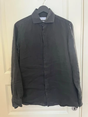 Svart linneskjorta med knappar - Stilren svart skjorta i linne med klassisk krage och knäppning framtill. Skjortan har långa ärmar och en avslappnad passform, perfekt för en trendig och enkel look.