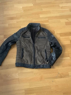 Brun och svart skinnjacka från rockandblue - Snygg brun och svart skinnjacka med dragkedja framtill och fyra fickor med dragkedja. Jackan har svarta skinnärmar och detaljer på axlar och nederkant. Modern passform och cool kontrast mellan material och färger.