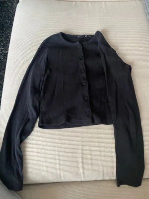 Svart ribbad kofta från H&M - Svart, croppad kofta från H&M med ribbad struktur och rund halsringning. Koftan har långa ärmar och knäpps med svarta knappar framtill. Perfekt att slänga över en topp eller klänning för en enkel och stilren look.