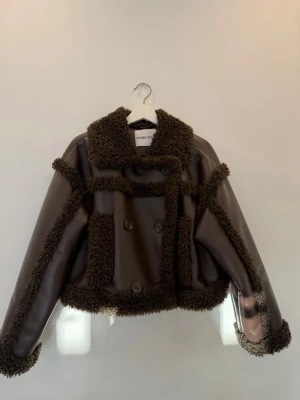 Brun teddyjacka från Stand Studio - Säljer en brun teddyjacka från Stand Studio med markerade sömmar och detaljer i mjuk teddy. Jackan har dubbelknäppning, stor krage och två framfickor. Materialet är en mix av fejkläder och teddy, perfekt för kalla dagar.