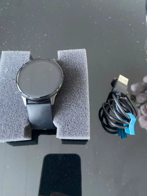 Svart smartklocka med silikonarmband - Snygg svart smartklocka med rund urtavla och matchande svart silikonarmband. Klockan har en stilren design med metallram och kommer med laddkabel. Perfekt för dig som vill ha koll på notiser och träning direkt på handleden.