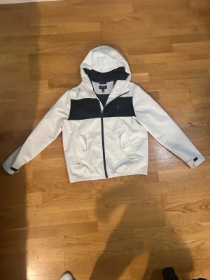 Vit vindjack från Polo Ralph Lauren - Snygg vit vintage vindjacka från Polo Ralph Lauren med marinblå panel över bröstet och huva. Jackan har dragkedja framtill, två fickor och justerbara muddar vid ärmslut. Klassisk röd Polo-logga på bröstet och sportig look som passar perfekt till kommande våren!