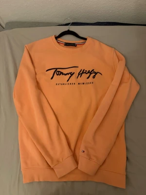 Persikofärgad s tröja Tommy Hilfiger - Persikofärgad s tröja från Tommy Hilfiger med svart broderad logga och texten 'Established MCMLXXXV' på bröstet. Tröjan har rund halsringning, långa ärmar och ribbade muddar vid ärmslut och nederkant. Liten flagglogga på ärmen. Jätte fin skick och knappt tecken på användning. Stolen är L så sitter bra ifall du är 180 och uppåt.