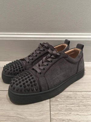 Gråa Christian Louboutin sneakers - Snygga gråa sneakers från Christian Louboutin i mocka med ikoniska nitar på tån och klassisk röd sula. Skorna har snörning och en stilren design som sticker ut med sina detaljer. Perfekta för dig som vill ha något unikt och trendigt. Skorna kommer inte komma i boxen och eftersom att den är sönder undertill