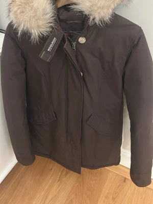 Brun parkas med fuskpäls från Woolrich - Mörkbrun parkas från Woolrich med stor huva och fluffig fuskpäls. Jackan har två snedställda fickor framtill, dragkedja och knappar. Perfekt för kalla dagar med sin varma och stilrena design.