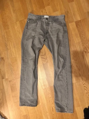 Grå raka jeans från Jack and Jones - 32/32 i storlek. Säljer ett par grå jeans med rak passform och klassiska bakfickor. Jeansen har en enkel design utan slitningar eller dekorationer och är perfekta för en avslappnad stil. Materialet är denim och färgen är en ljusgrå ton.