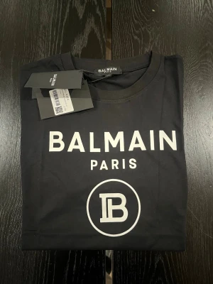 Svart Balmain Paris t-shirt - Snygg svart t-shirt från Balmain Paris med vit logotyptryck framtill. Klassisk rund halsringning och korta ärmar. Perfekt för dig som vill ha en stilren och trendig look med exklusiv känsla. Gjord i mjuk bomull för maximal komfort.