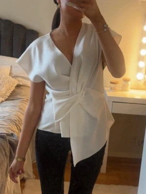 Vit blus  - Stilren vit omlottblus med korta, vida ärmar och snyggt knytband i midjan. Blusen har djup V-ringning och ett elegant fall. Perfekt för dig som gillar minimalistisk och modern stil. Materialet ser ut att vara ett mjukt, vävt tyg. Från Zara, Stl xs💕endast testad, som ny! 
