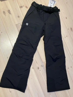 Svarta täckbyxor från 8848 Altitude storlek 150 - Svarta täckbyxor från 8848 Altitude med raka ben, justerbart bälte i midjan och två fickor framtill. Perfekta för vinterns alla äventyr och håller dig varm och torr. Snygg och stilren design med diskret logga på låret. Beninnerlängd 68 cm. Modell Inca, nya med lappar. Storlek 150.