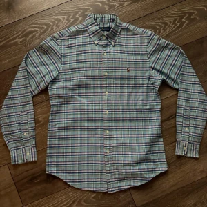 Ralph lauren skjorta - Klassisk långärmad skjorta från Ralph lauren med rutigt mönster i blått, grönt, vitt och rött. Skjortan har button-down krage, bröstficka med broderad logga och vita knappar. Tillverkad i mjuk bomull för en bekväm känsla.