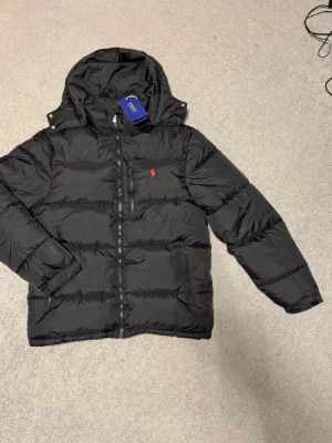 Svart dunjacka från Polo Ralph Lauren - Svart dunjacka med huva från Polo Ralph Lauren. Jackan har dragkedja framtill, två sidofickor och den klassiska röda loggan broderad på bröstet. Perfekt för kalla vinterdagar och riktigt snygg streetstil.