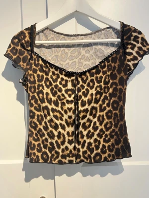 Leopardmönstrad topp med spetskant - Snygg topp med leopardmönster och svarta spetsdetaljer längs urringningen. Modellen har korta ärmar och en figurnära passform. Perfekt för dig som vill sticka ut med ett djurmönstrat plagg.