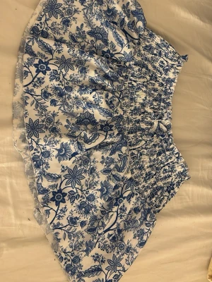 Vit och blå blommig kortkjol - Supersöt kortkjol med blått blommönster på vit botten. Kjolen har smock i midjan för skön passform och är gjord i ett lätt och luftigt material, perfekt för varma dagar. Passar perfekt till en enkel topp eller linne.