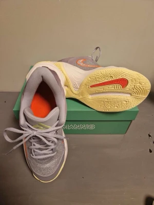 Nike Giannis grå basketskor - Snygga basketskor från Nike Giannis-serien i grått med orange detaljer och limegröna accenter. Ovandelen är i mesh och syntet för bra andningsförmåga och komfort. Sulan är ljusgul med röd Nike-logga undertill. Perfekta för basket eller streetstyle.