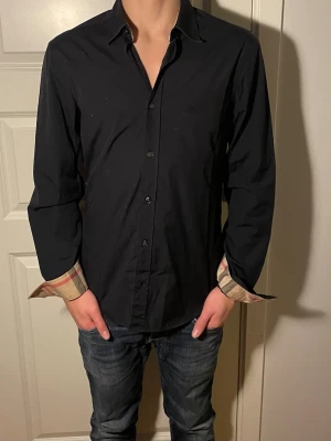 Burberry skjorta  - Säljer en svart skjorta från Burberry med klassiskt rutigt mönster på insidan av manschetterna och kragen. Skjortan har knappar framtill och lång ärm. Perfekt för dig som gillar stilrena plagg med en twist.